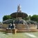 A fountain with statues and jets of water near trees and onlookers